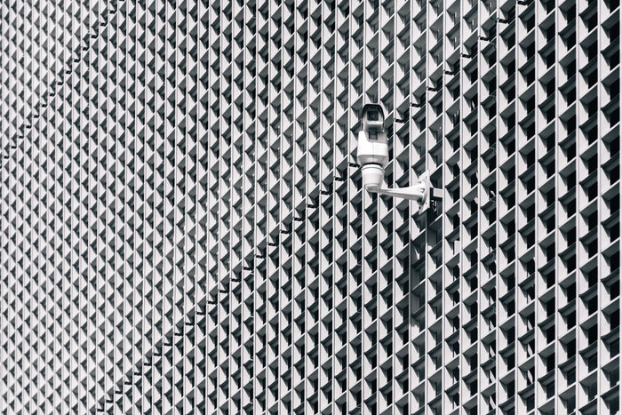 This image features a surveillance camera installed on the exterior of a contemporary building with a repeating geometric window pattern. The high-contrast architectural design emphasizes security and monitoring in urban environments, highlighting the role of CCTV systems in maintaining safety and surveillance across commercial or public spaces.